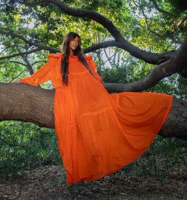 Brooke DeVard poses in a flowing orange dress along a tree branch, with Naked Beauty resting beside her on the same branch.